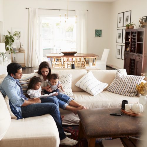 Young Hispanic family sitting on sofa reading a book together in the living room, seen from doorway